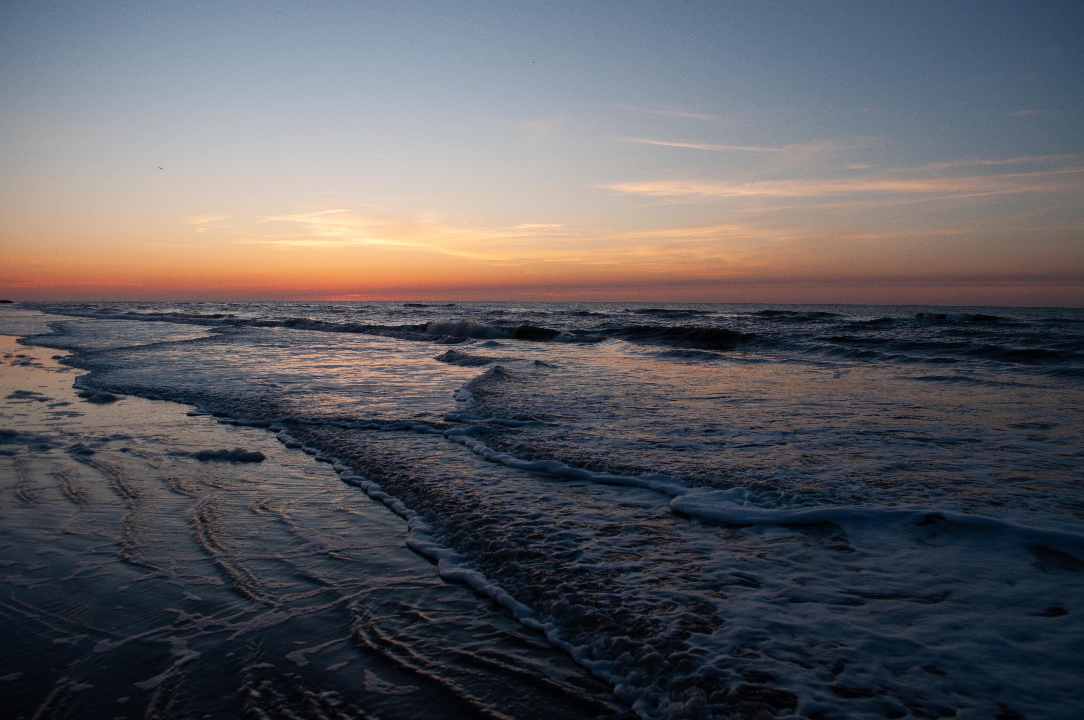 A quiet shoreline at dusk, waves catching the last warm light of a fading sunset over the horizon.
